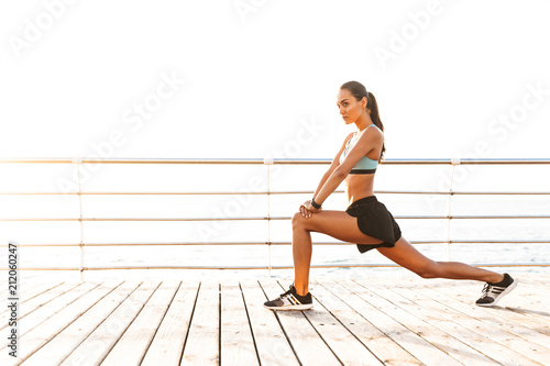 Profile photo of slim healthy woman 20s in tracksuit squatting, and stretching legs on boardwalk at seaside