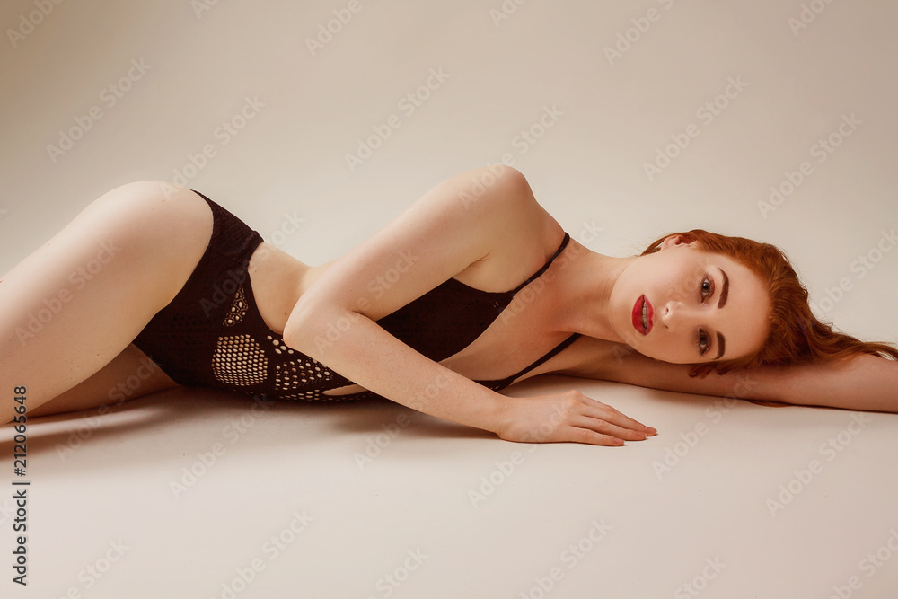 Fototapeta premium Model shoot in studio. Skinny girl with red long hair posing in the black knitted swimsuit bikini lying on the floor in warm tones