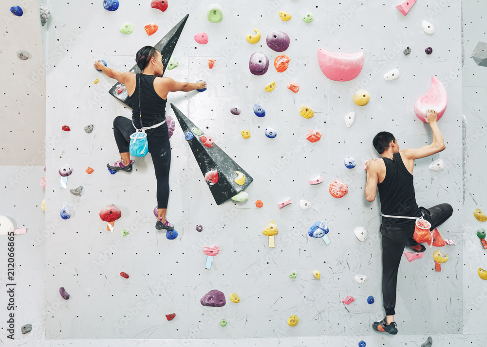 Back view of sportive people in black outfits ascending artificial boulders on clambering wall in modern gym Back view of sportive people in black outfits ascending artificial boulders on clambering wall in modern gym