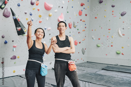 Young Asian man and woman with sack of talk powder on hips standing in climbing gym and looking up in excitement