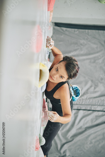 From above shot of serious ethnic woman clambering wall with boulders in gym and looking at camera