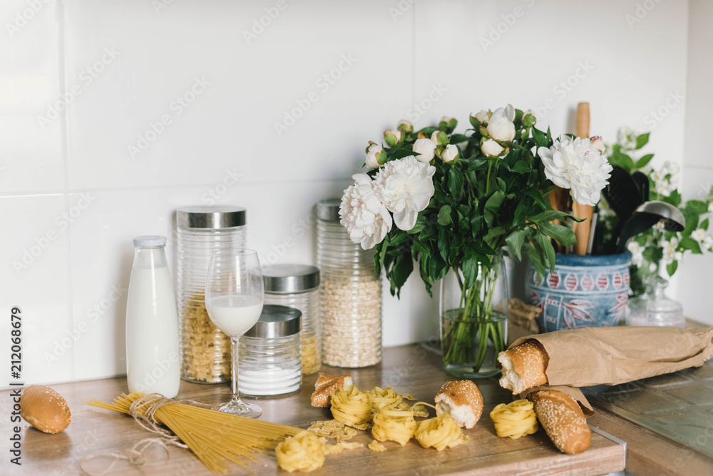White tiles wall modern kitchen with chopping board,flowers,knifes,pasta,bread. White tiles wall modern kitchen with white top background and ingredients - obrazy, fototapety, plakaty 