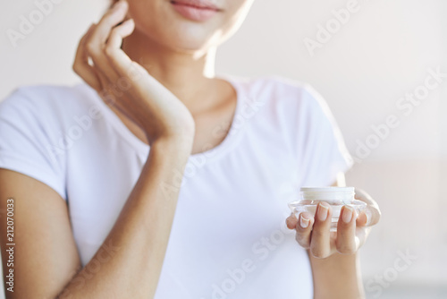 Crop female in white t-shirt taking care of skin putting cream on face on light blur background