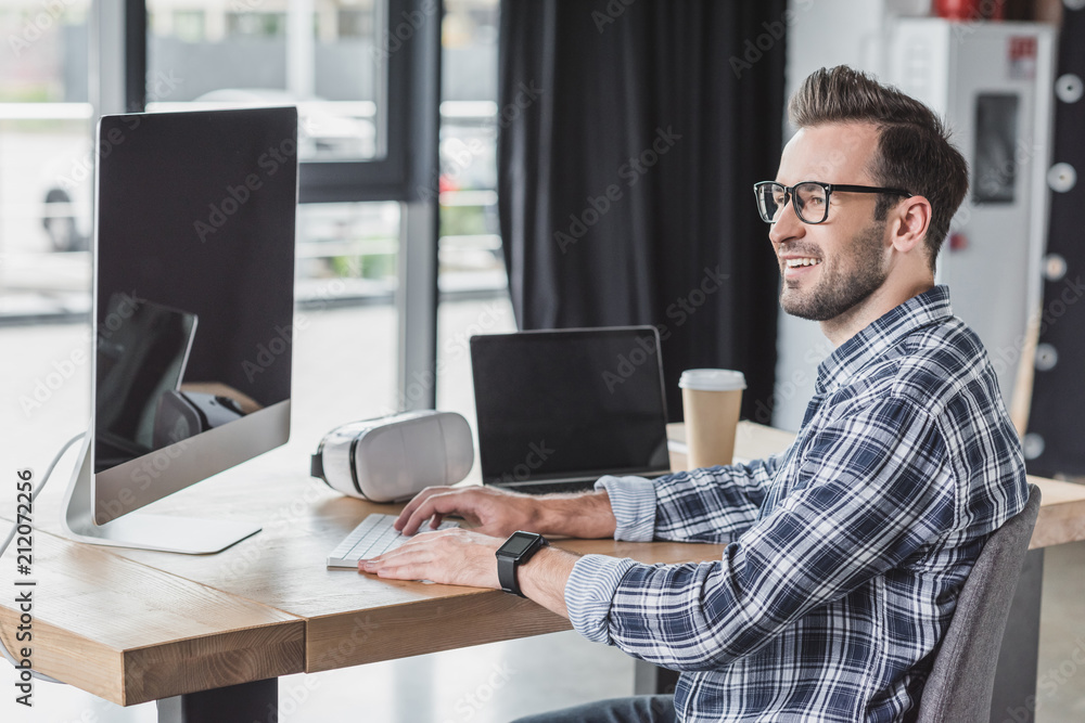 smiling young man in eyeglasses using desktop computer and laptop at workplace smiling young man in eyeglasses using desktop computer and laptop at workplace