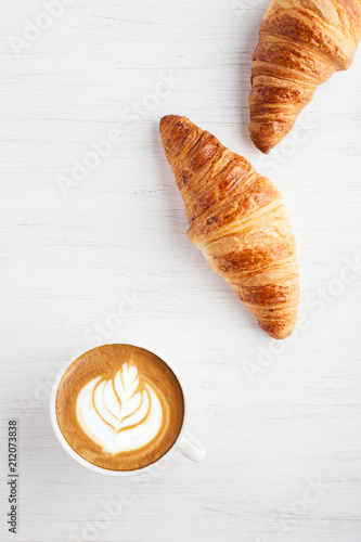 A cup of cappuccino with latte art and two croissants on white wooden table. Traditional french breakfast. Top view.