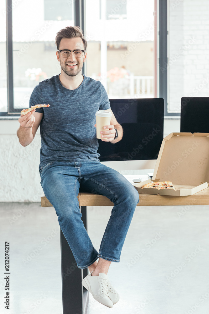 handsome smiling young programmer in eyeglasses holding paper cup and eating pizza at workplace