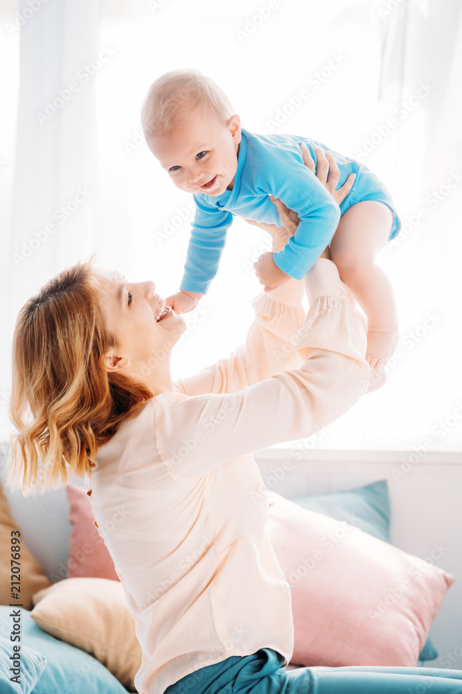 side view of mother raising laughing little child while sitting on bed at home side view of mother raising laughing little child while sitting on bed at home
