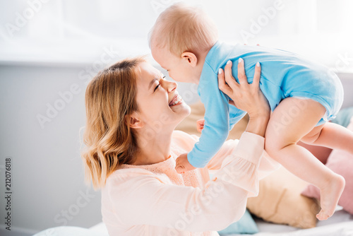 smiling mother carrying happy baby while sitting on bed