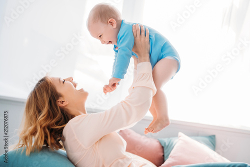 side view of happy mother raising laughing little child while sitting on bed at home