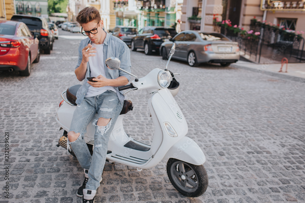 Handsome guy is sitting on motorcycle and holding phone in hand. He is looking at it. Theere is a cigarette in other hand. He is smoking.