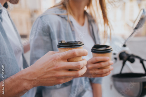 Close up of guy's and girl's hands holding cups of coffee. This poeple are sitting on motorcylcle and having some rest.