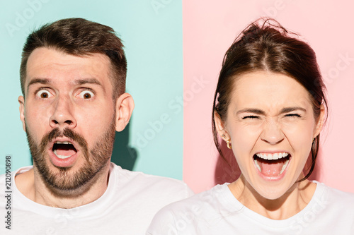 Closeup portrait of young couple, man, woman. One being excited happy smiling, other serious, concerned, unhappy on pink and blue background. Emotion contrasts