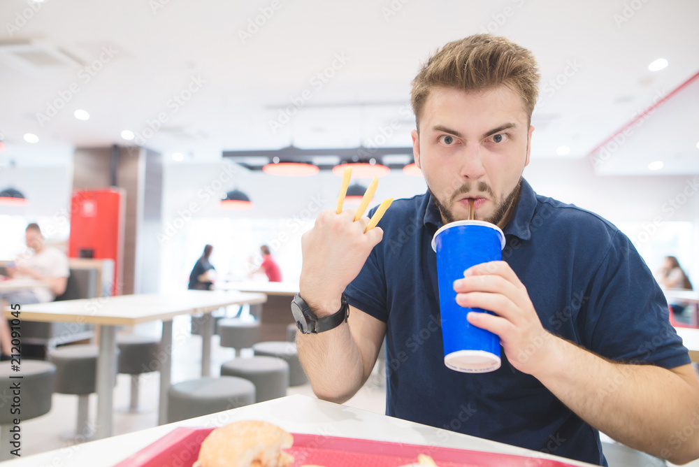 Funny man with french fries and a glass of cool drink sits in a restaurant and looks at the camera. Emotional man drinks and eats French fries in a restaurant. Funny man with french fries and a glass of cool drink sits in a restaurant and looks at the camera. Emotional man drinks and eats French fries in a restaurant.