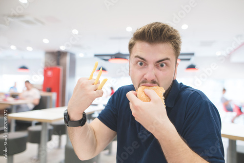 Portrait of a funny emotional man with French fries instead of fingers who eats a burger in a fast-food restaurant and looks at the camera. Fast Food Concept