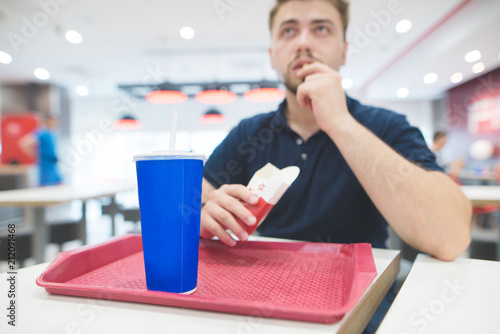 Man eats quick food from the red tray at the restaurant at the table. A blue glass with a cool sweet drink on the background of a man who eats French fries. Focus on the glass. Fast Food Concept. photo