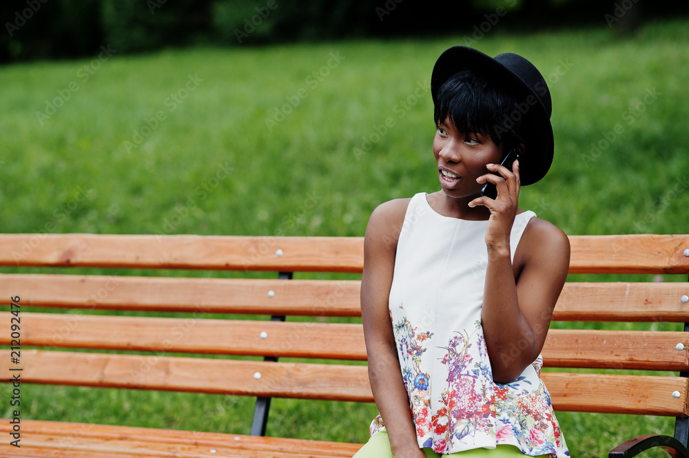 Naklejka premium Amazing african american model woman in green pants and black hat posed on bench at park and speaking on mobile phone.