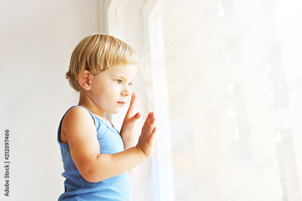 Fototapeta premium Child missing his mother concept. Little cute blond three year old boy sitting on windowsill touching window glass, looking sad. Kid in blue tank top playing alone. Background, close up, copy space.