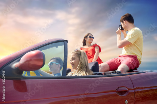leisure, road trip, travel, summer holidays and people concept - happy friends driving in convertible car and taking picture by film camera over sky background © Syda Productions