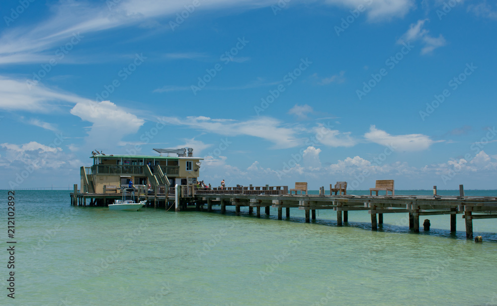 Old wooden fishing pier with building, people, tropical beach, blue sky.