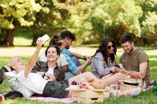 friendship, leisure, technology and people concept - group of friends with smartphones and food chilling on picnic blanket at summer park