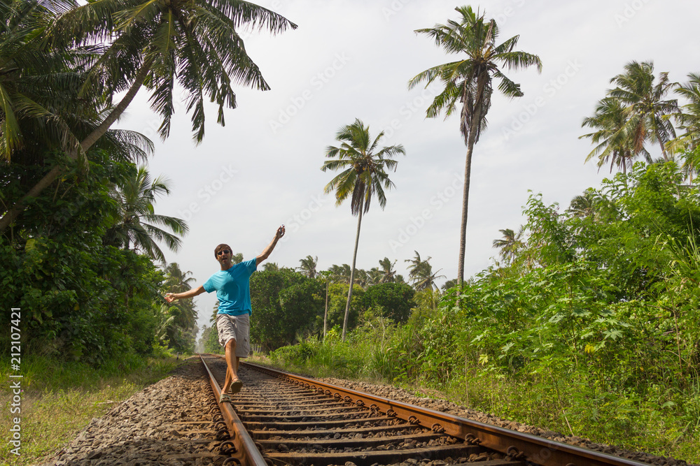 Young man walking on railroad track while listening to music with headphones on his smartphone in rural area in Bentota, Sri Lanka. Reckless, temerarious concept