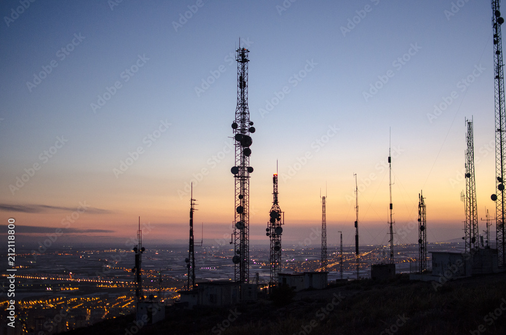 communication tower. cell, radio and television antennas on top of a mountain and below a lit coastal village - obrazy, fototapety, plakaty 