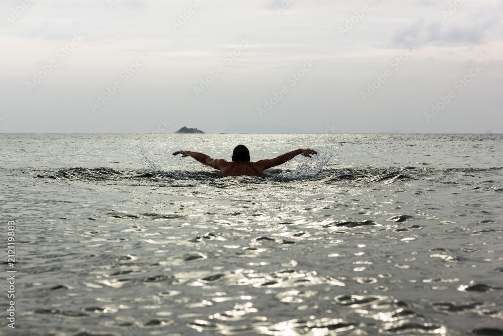 Silhouette of young man swimming away on butterfly stroke technique towards a small island on the background at sunset in Koh Phangan, Thailand. Hard to reach, destination challenge, effort concepts
