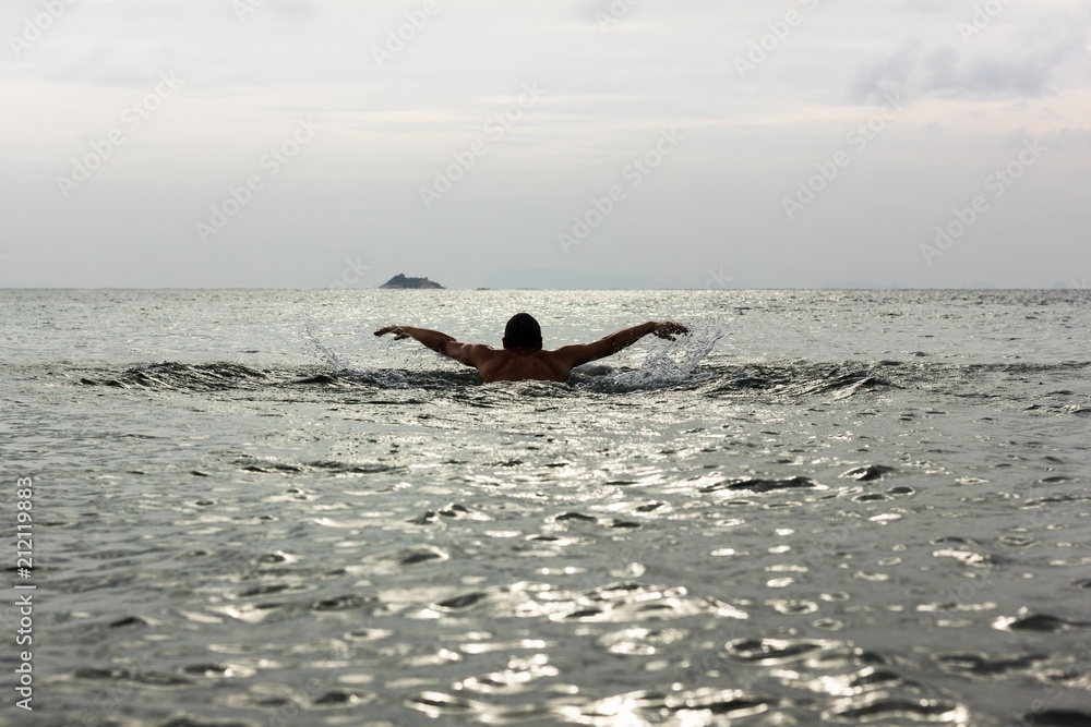 Naklejka premium Silhouette of young man swimming away on butterfly stroke technique towards a small island on the background at sunset in Koh Phangan, Thailand. Hard to reach, destination challenge, effort concepts