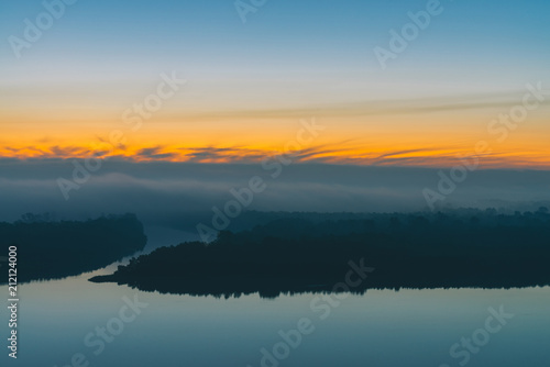 Early blue sky reflected in river water. Riverbank with forest under predawn sky. Yellow stripe in picturesque sky. Fog hid trees on island. Mystical morning atmospheric landscape of majestic nature. © Daniil