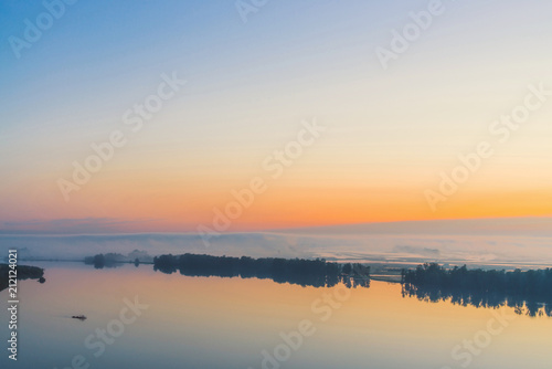 Broad mystical river flows along diagonal shore with silhouette of trees and thick fog. Gold glow in predawn sky. Calm morning atmospheric landscape of majestic nature in warm tones.