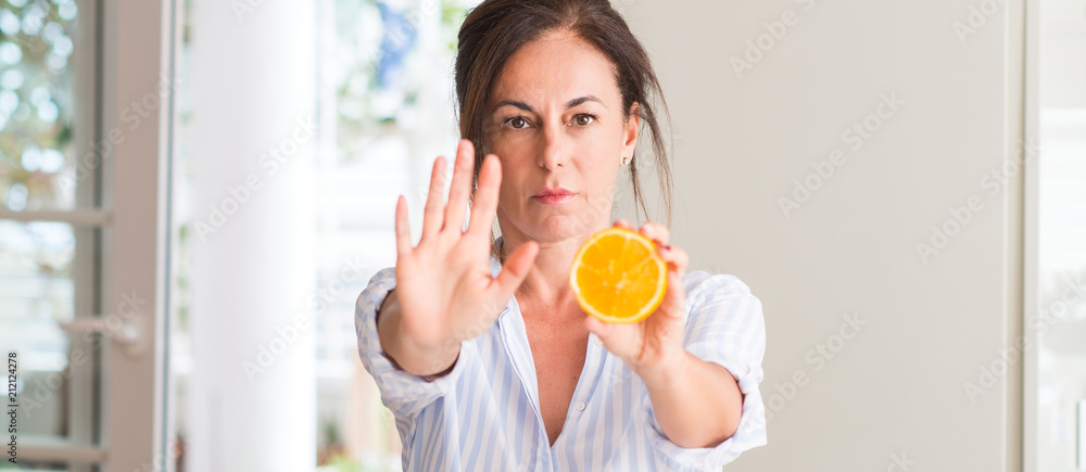 Middle aged woman holding orange fruit with open hand doing stop sign with serious and confident expression, defense gesture Middle aged woman holding orange fruit with open hand doing stop sign with serious and confident expression, defense gesture