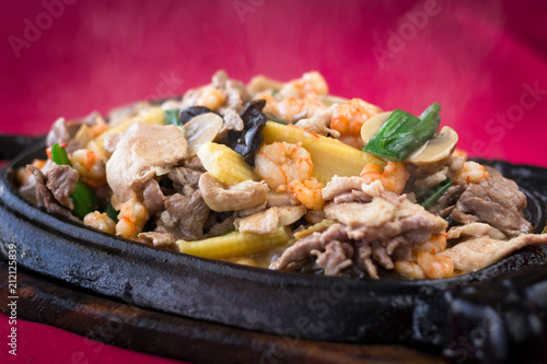 Close up on smokey meat sizzler with prawns, baby corn and mushrooms on iron black plate with wooden base on red tablecloth background. Traditional Chinese hot menu dish