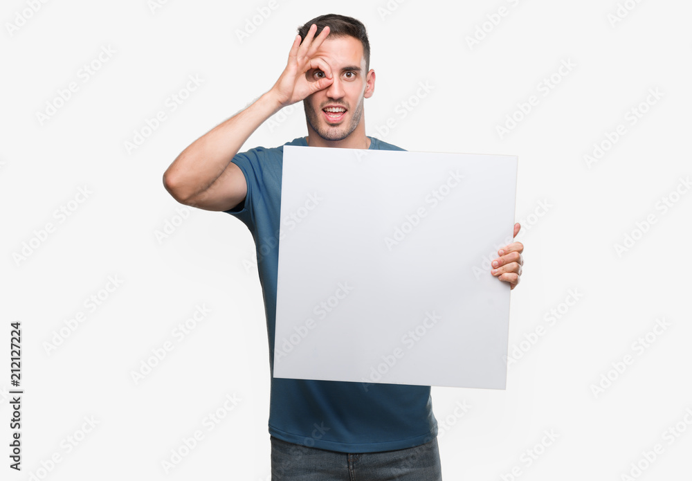 Handsome young man holding advertising banner with happy face smiling doing ok sign with hand on eye looking through fingers Handsome young man holding advertising banner with happy face smiling doing ok sign with hand on eye looking through fingers