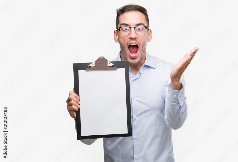 Handsome young business man showing a clipboard very happy and excited, winner expression celebrating victory screaming with big smile and raised hands Handsome young business man showing a clipboard very happy and excited, winner expression celebrating victory screaming with big smile and raised hands