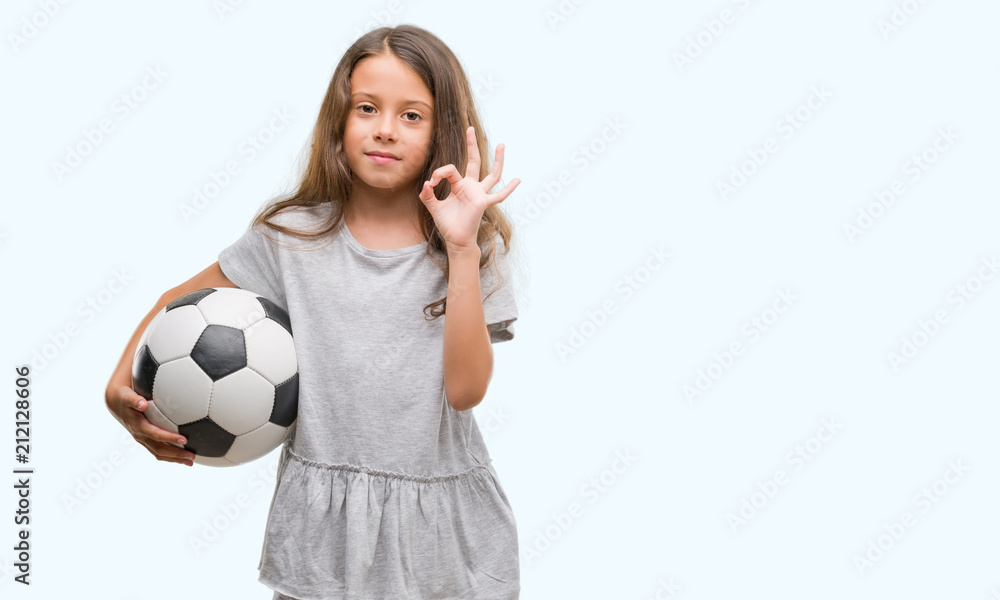 Brunette hispanic girl holding soccer football ball doing ok sign with fingers, excellent symbol Brunette hispanic girl holding soccer football ball doing ok sign with fingers, excellent symbol