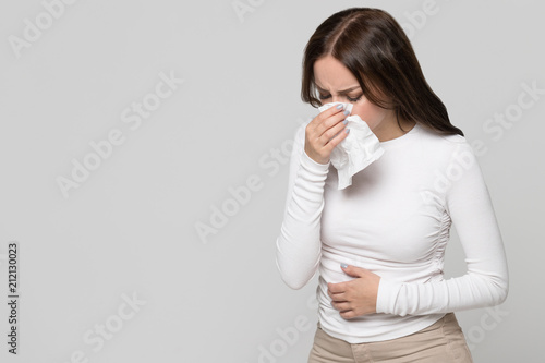 Studio portrait of female with napkin sneezing, experiences allergy symptom, isolated on grey background. Rhinitis, runny nose