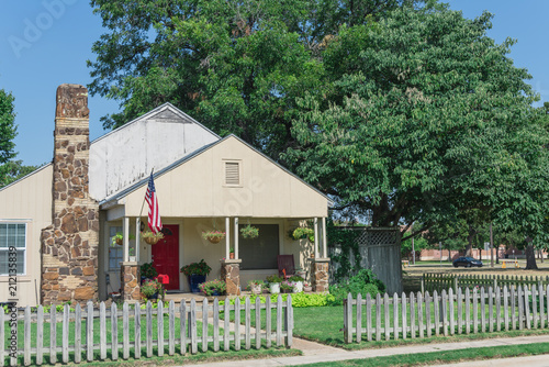 Modest house with chimney in historic downtown district of Irving, Texas, USA. Classic wooden fence with well-groomed landscape, haning flower pots, big tree and proudly American flag waving