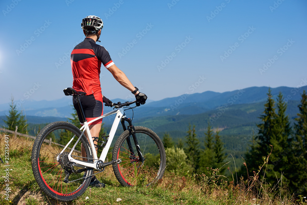 Back view of athletic professional bicyclist stopped on top of hill to enjoy beautiful view of distant Carpathian mountains on blue summer sky background. Active lifestyle and extreme sport concept.