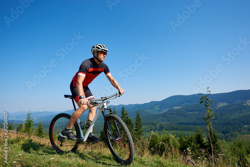 Young athletic tourist cyclist in helmet, sunglasses and full equipment riding bike down grassy hill on distant mountains and blue summer sky background. Active lifestyle and extreme sport concept.