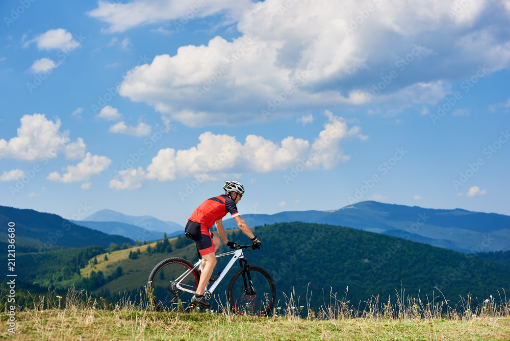 Obraz premium Professional sportsman cyclist in sportswear and helmet riding cross country bike in high grassy hill. Beautiful mountains view and cloudy sky on background. Active lifestyle and extreme sport concept