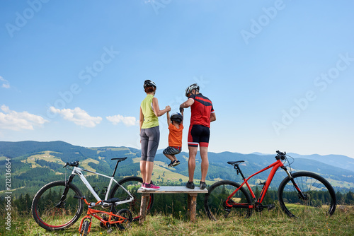 Young family tourists cyclists, mother, father and child, resting with bikes, sitting on wooden bench on grassy hill on distant mountains view background. Active lifestyle and happy relations concept.