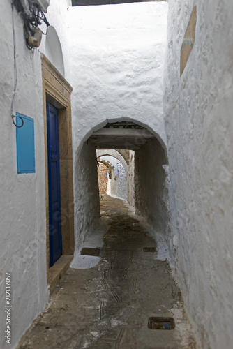 A view of a narrow street with arch and wooden windows and doors with white wall stone architecture of the island Patmos, Greece © CoolimagesCo