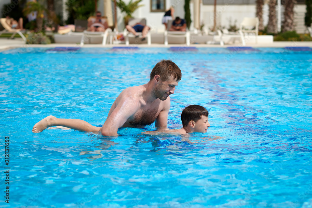 Cute caucasian boy is obtaining swimming skills. His father is helping him.
