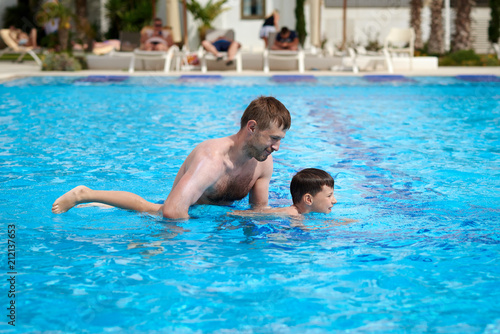 Cute caucasian boy is obtaining swimming skills. His father is helping him.