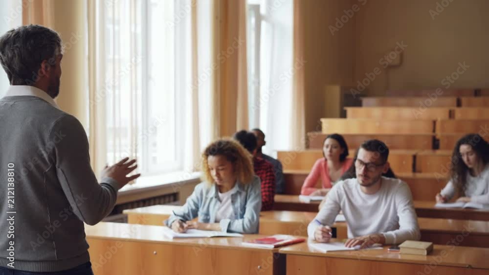 Male teacher is explaining new information to multi-ethnic group of students who are writing in notebooks taking notes. Profession, education and youth concept. Male teacher is explaining new information to multi-ethnic group of students who are writing in notebooks taking notes. Profession, education and youth concept.
