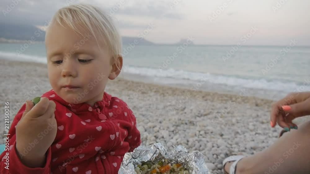 Close-up of two years old baby eating green olive from a foil tray on the beach. Child and mom on the sunset beach Close-up of two years old baby eating green olive from a foil tray on the beach. Child and mom on the sunset beach