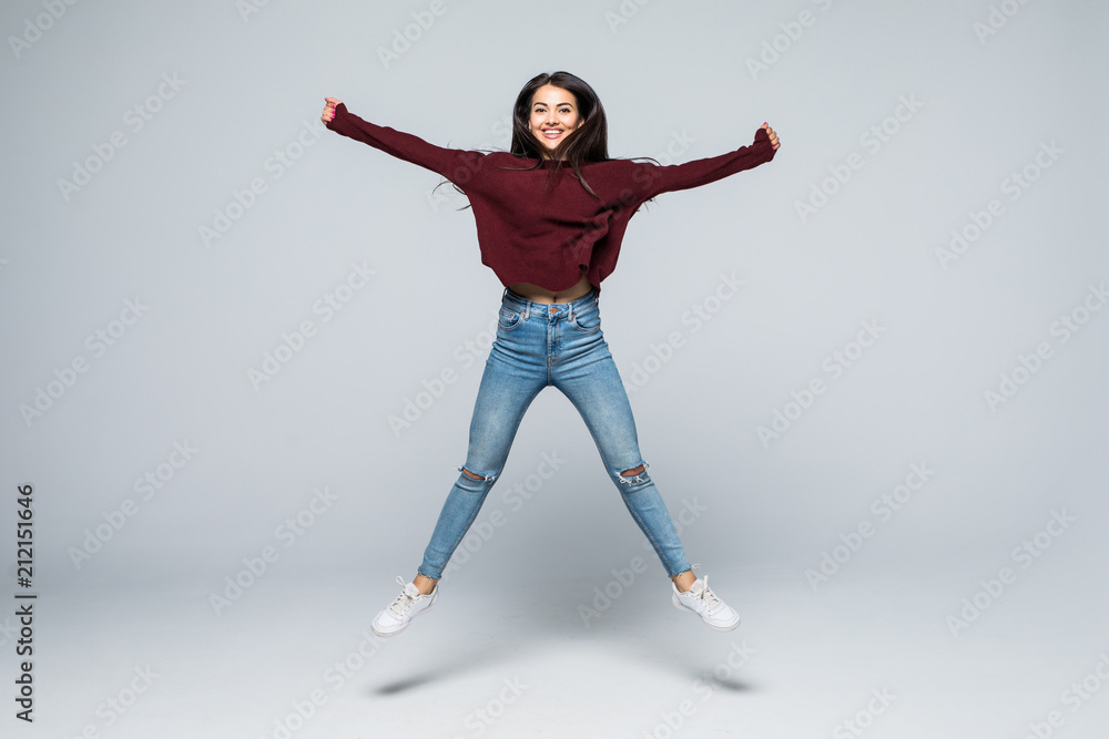 Fototapeta premium Pełnometrażowy portret ładnej, dziewczęcej kokietki, uroczej, nowoczesnej, stylowej dziewczyny w dżinsowym stroju, spodniach, skaczącej w powietrzu z uniesioną nogą, patrząc na kamery, na białym tle na szarym tle