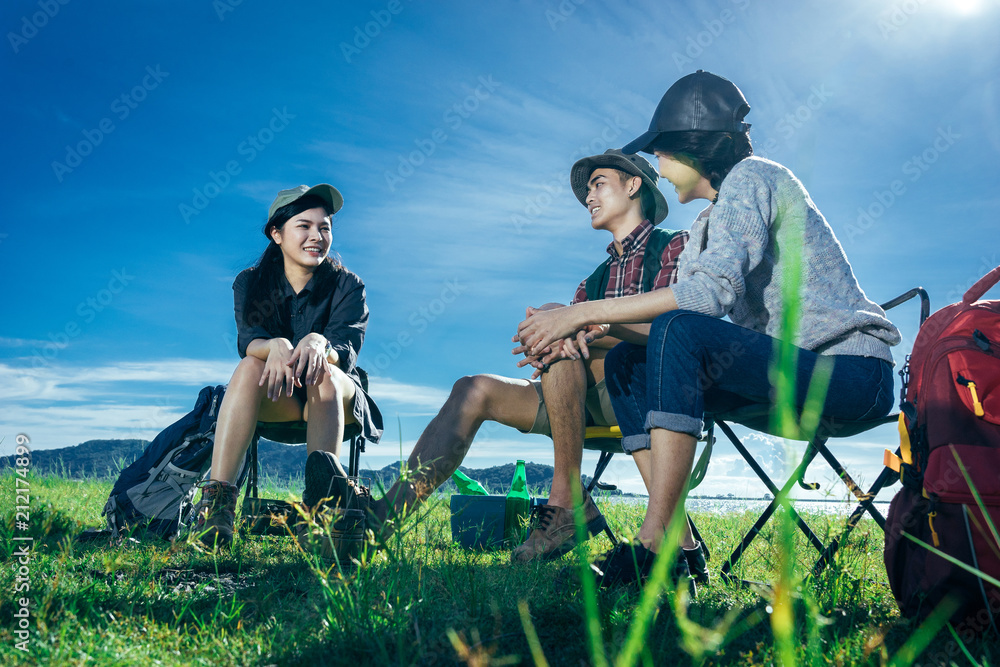 A  group of Asian friends sitting on chairs, drinking some beer and water together outside the tent near the fire while they has camping on Weekend holiday.
