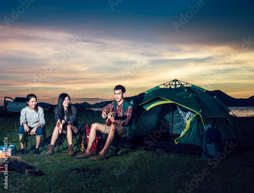 A cropped photo of  group of Asian friends sitting on chairs, singing, playing a guitar and drinking some beer and water together outside the tent while they has camping on Weekend holiday.