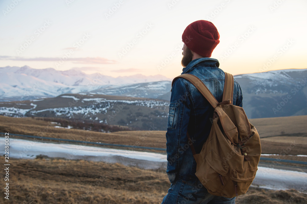 Bearded tourist hipster man in a hat with a backpack stand back on a roadside bump and watching the sunset against the background of a snow capped mountain